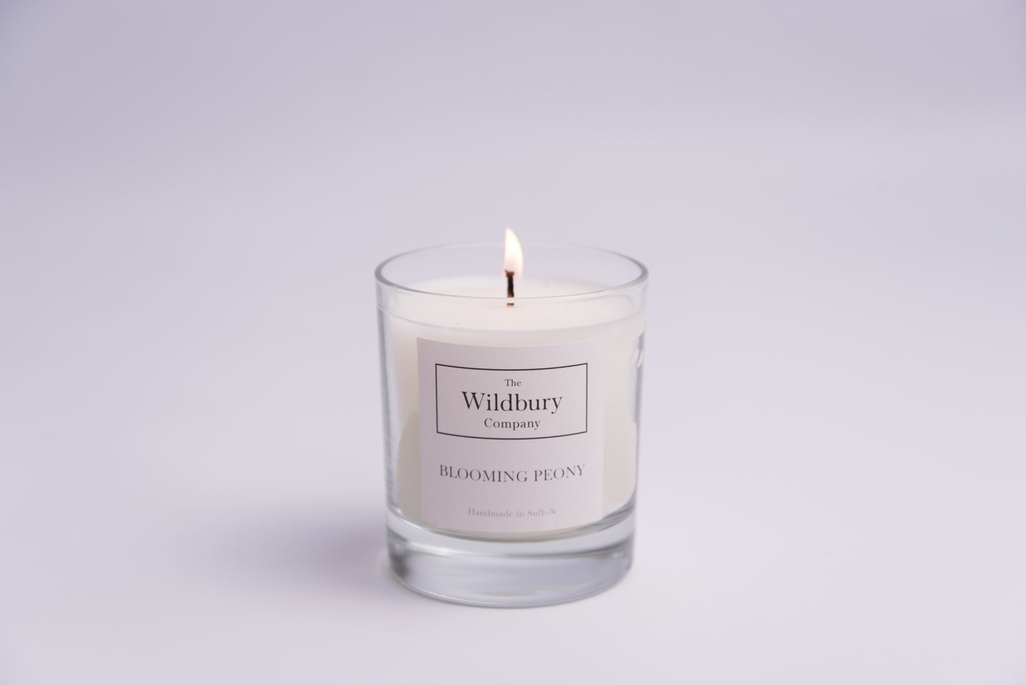The Wildbury Company Single Wick Signature Candle in Glass burning with flame and label on the glass.